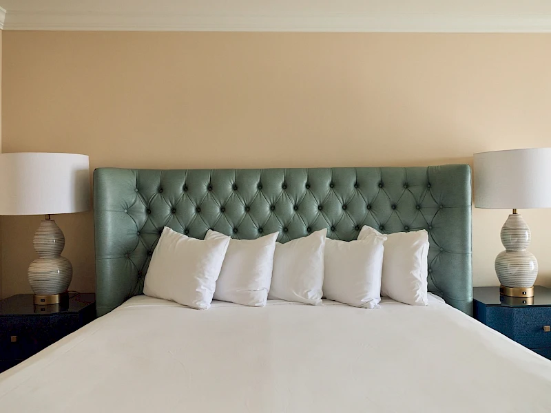 A neatly made bed with a tufted green headboard, five white pillows, matching nightstands, and two lamps flanking the bed.