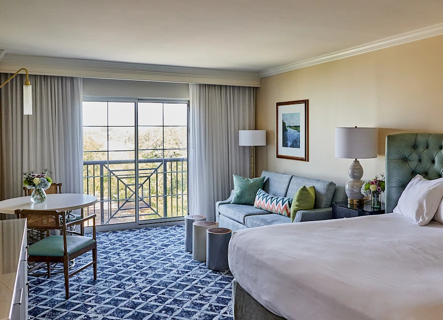 A bright hotel room with a king bed, tufted headboard, sofa, round table and chairs, a balcony with sliding doors, and soft decor in blue and beige tones.