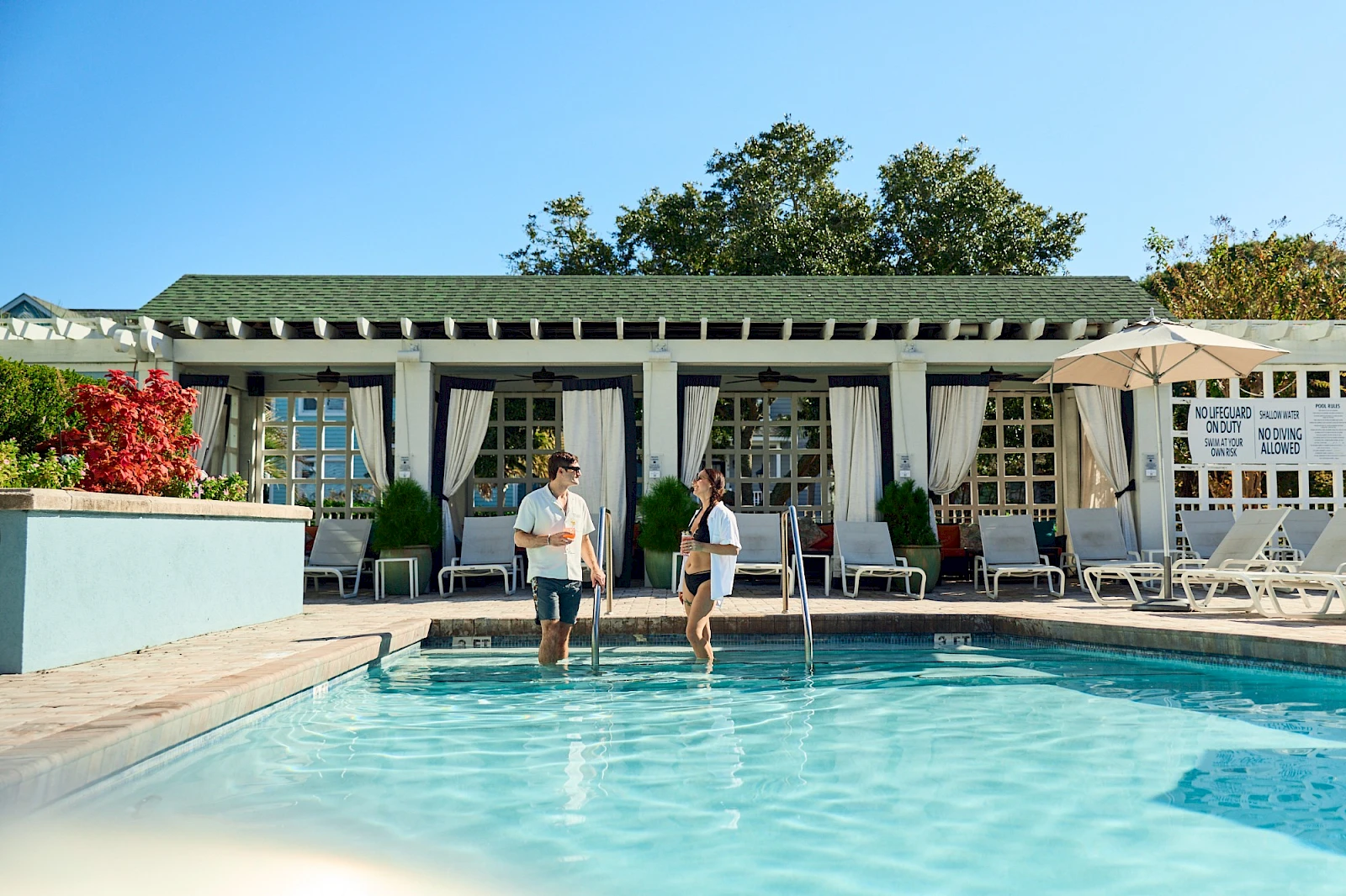 Two people walk from the pool through a sunny resort-style patio toward a white-clad building with lounge chairs and umbrellas, bright blue sky.