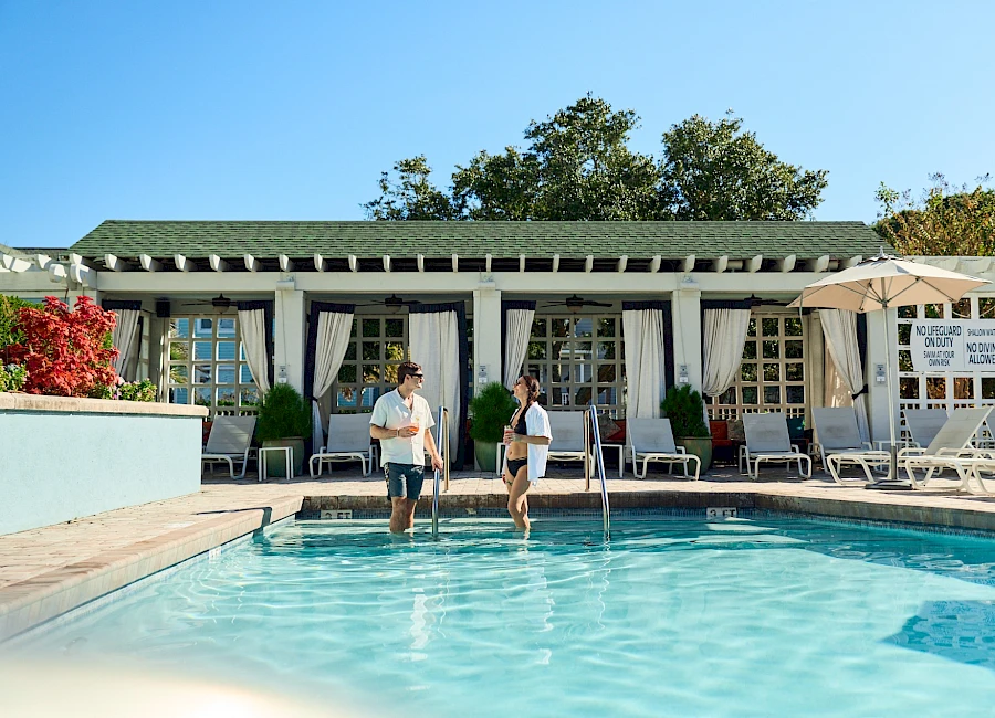 Two people walk from the pool through a sunny resort-style patio toward a white-clad building with lounge chairs and umbrellas, bright blue sky.