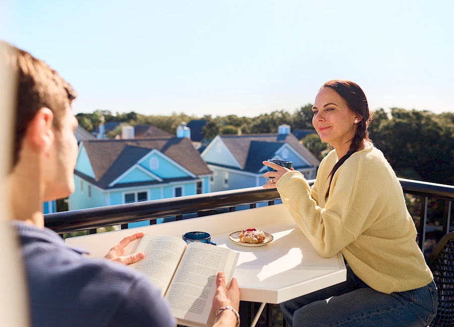 A couple sits on a balcony at a table with books and snacks, having a chat with a sunny neighborhood view behind them.