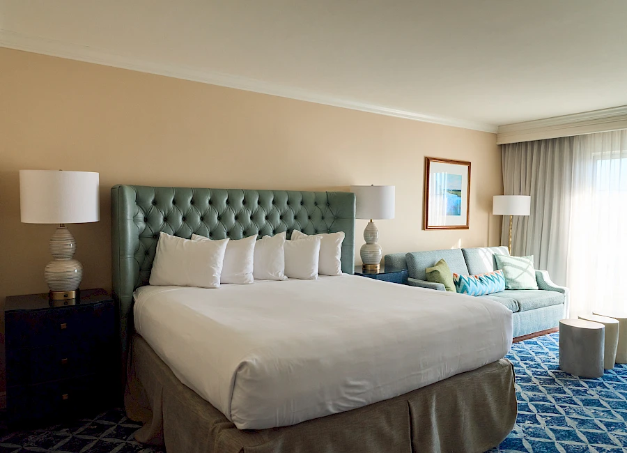A hotel room with a king bed, teal tufted headboard, two nightstands, lamps, a blue sofa, and a sunny window with sheer curtains.