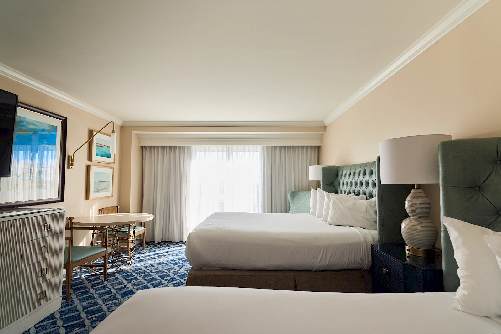 A bright hotel room with two queen beds, a blue tufted headboard, nightstand lamps, a desk, dresser, and a large window with sheer white curtains.