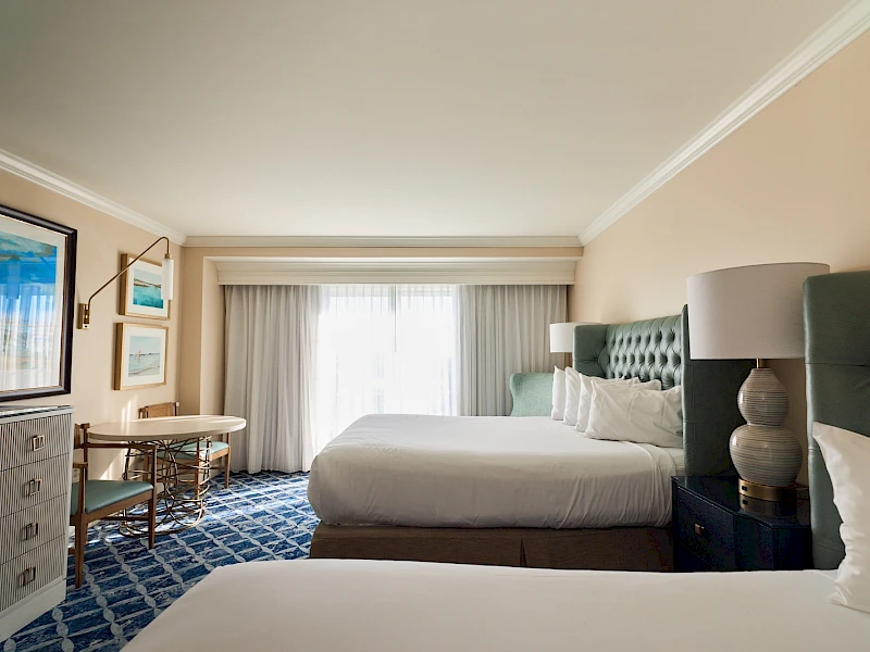 A bright hotel room with two queen beds, a blue tufted headboard, nightstand lamps, a desk, dresser, and a large window with sheer white curtains.