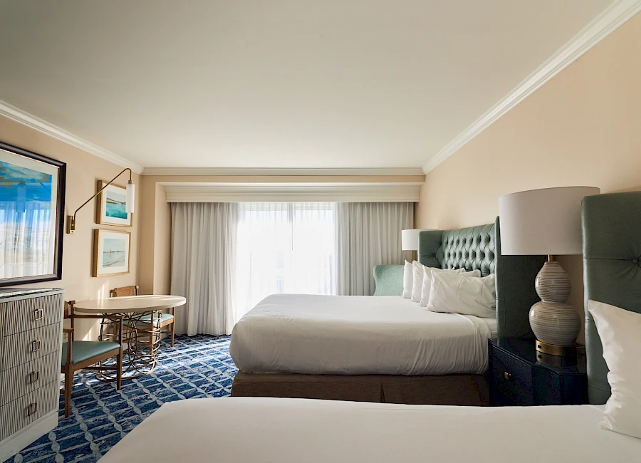 A bright hotel room with two queen beds, a blue tufted headboard, nightstand lamps, a desk, dresser, and a large window with sheer white curtains.