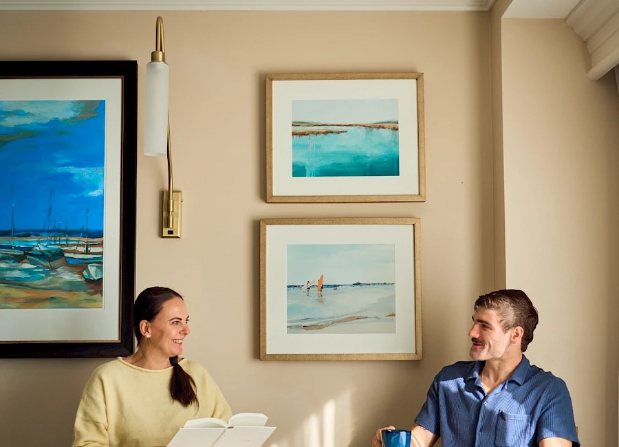 Two people sit at a small round table in a bright dining nook, chatting over coffee and pastries, with coastal artwork on the beige walls.