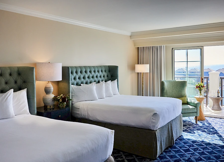 Two queen beds in a hotel room with teal tufted headboards, nightstands, lamps, a seating chair, and a balcony with ocean view.