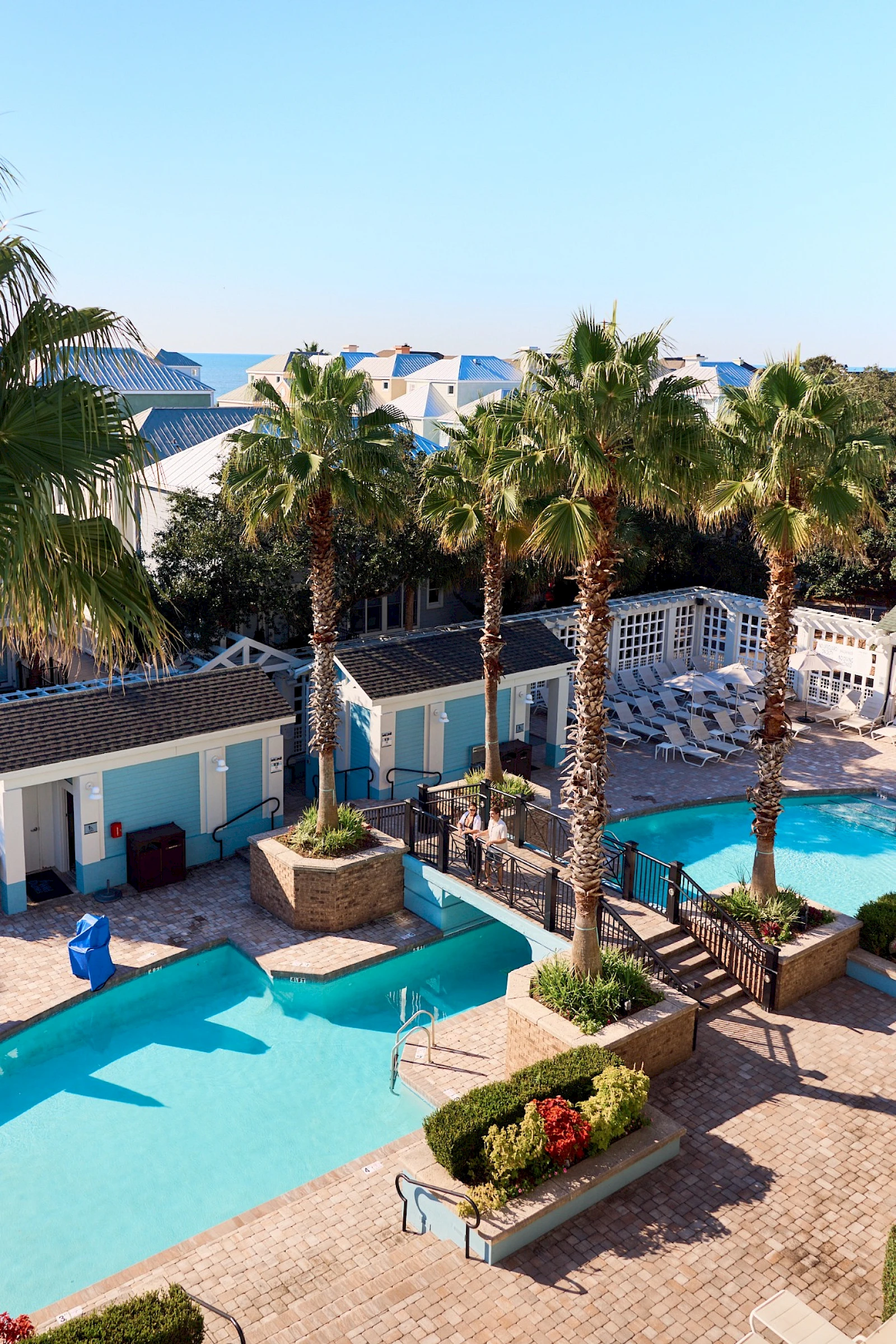 A sunny resort pool area with blue-tiled pools, palm trees, lounge seating, and small buildings with blue doors, all under a clear sky.