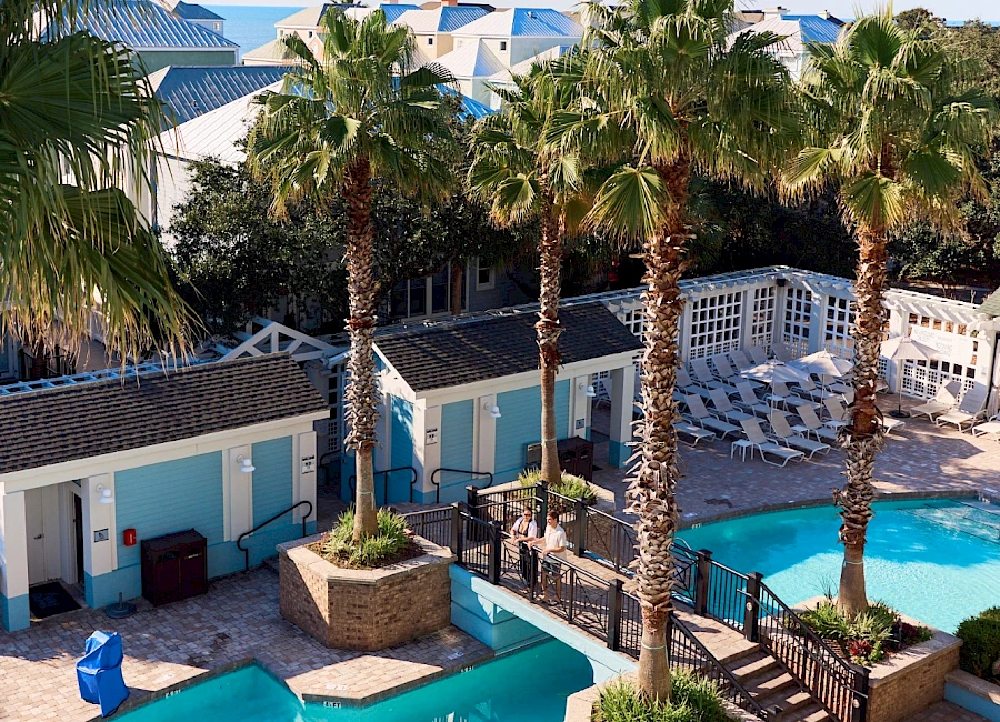 A sunny resort pool area with blue-tiled pools, palm trees, lounge seating, and small buildings with blue doors, all under a clear sky.