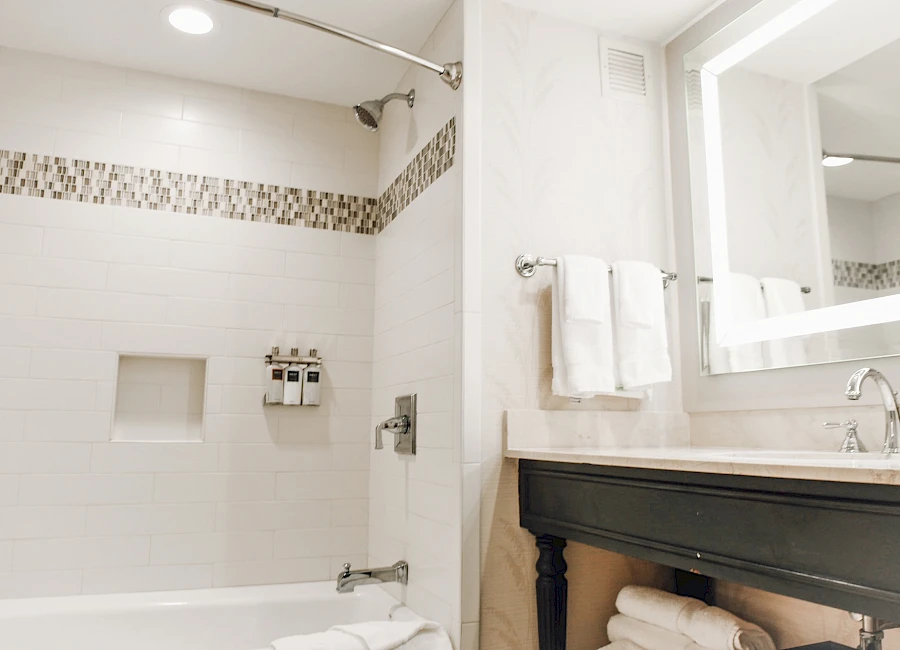 A clean, modern bathroom with a white tile shower, built-in niche, and a white tub; a dark vanity with a large mirror and folded towels.