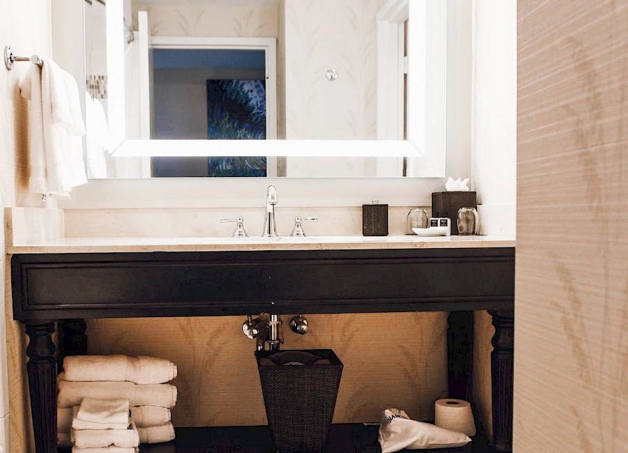 A bathroom vanity with a large illuminated mirror, towels on the shelves, and a small basket under the sink, all in a clean, modern setup.