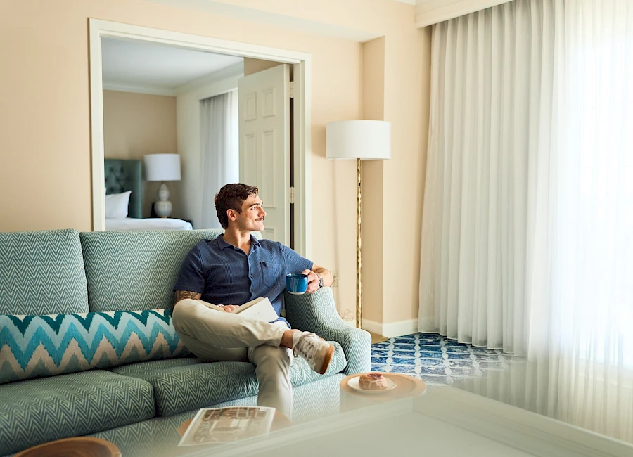 A man sits relaxed on a teal sofa in a bright hotel room, with a large mirror, floor lamp, and floor-to-ceiling sheer curtains by the window.