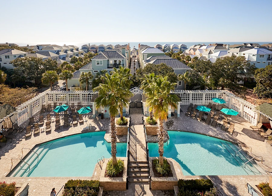 A sunny beachside community with a rectangular pool, palm trees, and lounge chairs, surrounded by white fences and cozy homes in the background.