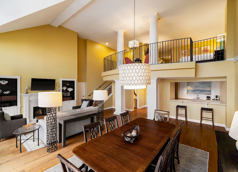 A spacious, two-story living area with a long wooden dining table, warm yellow walls, comfy seating, a mezzanine, and modern decor.
