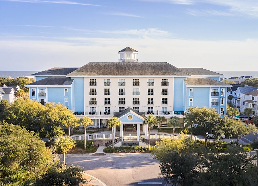 A blue seaside hotel with a central entrance, white trim, cupola on the roof, surrounded by green trees and a quiet street.
