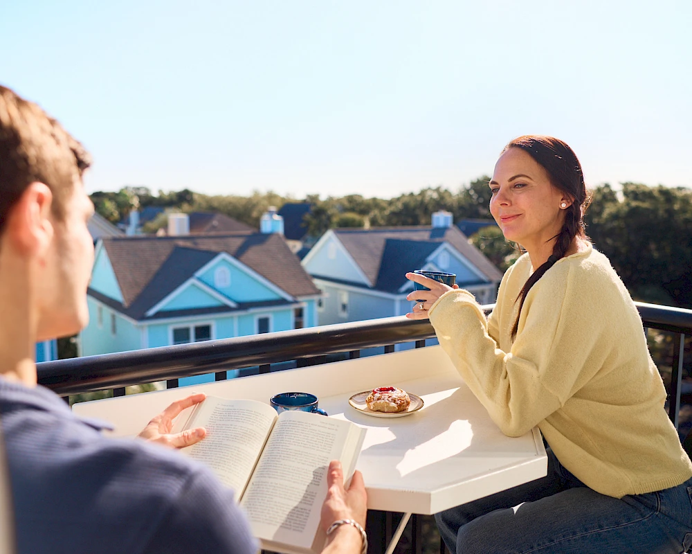 Two people dine on a balcony, enjoying coffee and a pastry while reading, with a sunny neighborhood backdrop.