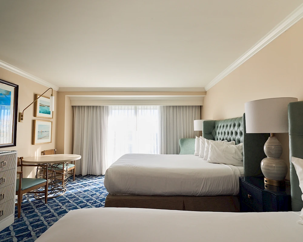 A hotel room with two queen beds, a patterned blue carpet, a desk and chair, wall lamps, a dresser, and a window with sheer curtains, bright and clean.