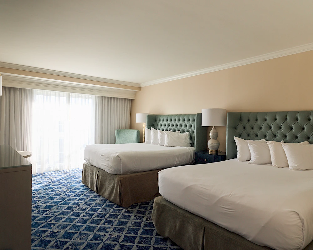 A hotel room with two queen beds, tufted headboards, nightstands, lamps, a chair, and a large window with curtains, plus a dresser—cozy and neat.