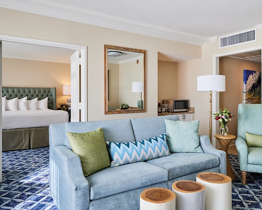A stylish hotel suite: living area with blue sofa, zigzag pillow, round ottomans, wood coffee table, and a glimpse of a bedroom with king bed.