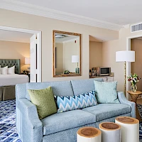 A stylish hotel suite: living area with blue sofa, zigzag pillow, round ottomans, wood coffee table, and a glimpse of a bedroom with king bed.