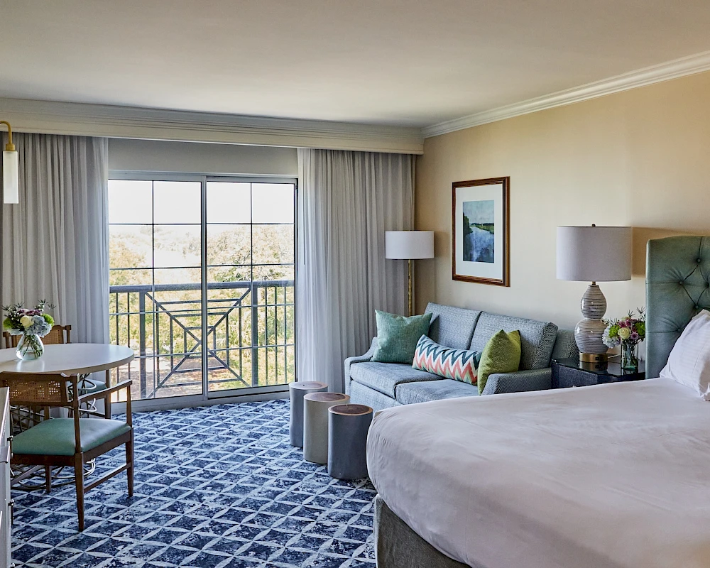 A hotel room with a king bed, teal headboard, sofa, round table, and mismatched cushions, plus a balcony view and blue patterned carpet.