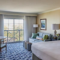 A hotel room with a king bed, teal headboard, sofa, round table, and mismatched cushions, plus a balcony view and blue patterned carpet.