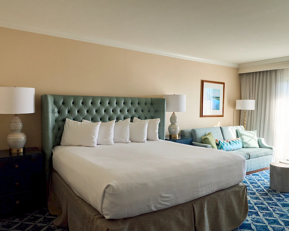 A hotel room with a large bed, green tufted headboard, two bedside lamps, a seating area with blue-green sofa, and a windowed balcony.