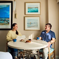 Two people sit at a round table chatting over coffee and pastries in a cozy cafe adorned with framed ocean art.