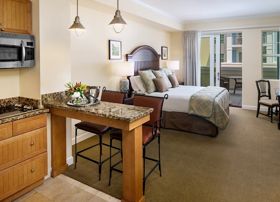 A cozy studio suite with a kitchen bar, granite counters, a bed, living area, and a balcony door, blending kitchenette, seating, and sleeping space.