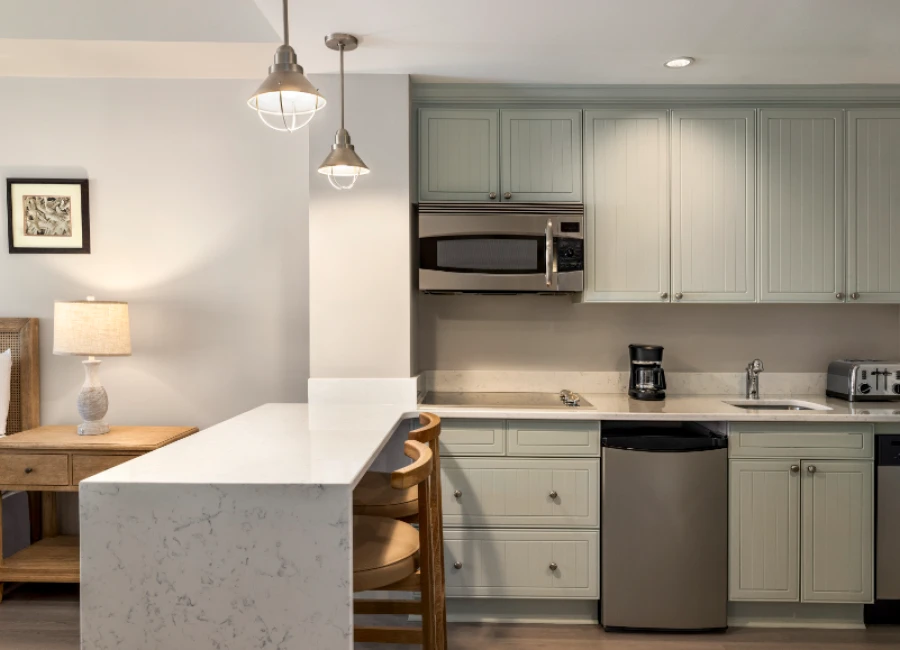 A modern kitchenette with light blue cabinets, stainless appliances, a white quartz island, two pendant lights, and a small dining bar with wooden stools.