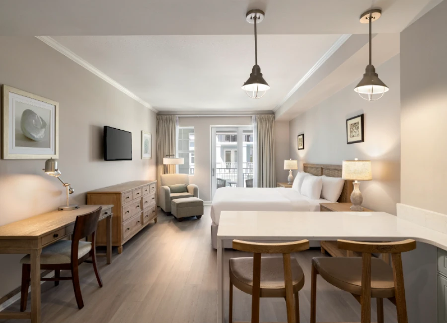 A bright hotel room with two queen beds, a seating area, desk, wall-mounted TV, and large windows; warm lamps and wooden furniture create a cozy, modern space.