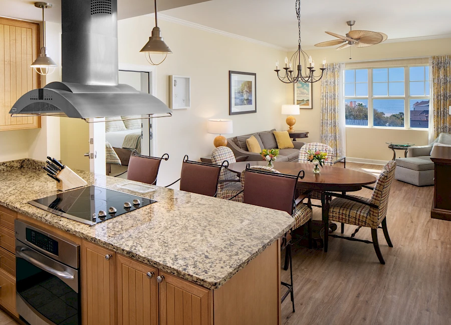 A bright open-concept kitchen and living room with a granite island, stainless cooktop, wooden cabinets, a dining table, beige walls, and a cozy seating area by large windows featuring a chandelier and ceiling fan.