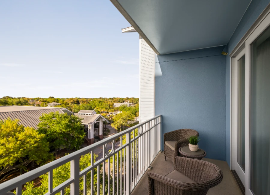 A balcony with wicker chairs and a small table overlooks a tree-filled street under a clear blue sky.