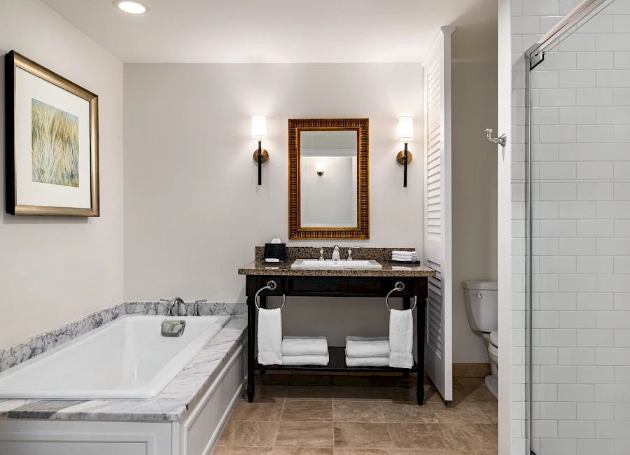 A modern hotel bathroom with a bathtub, a dark wooden vanity, a framed mirror, two wall sconces, and a glass-enclosed shower, all neatly arranged.