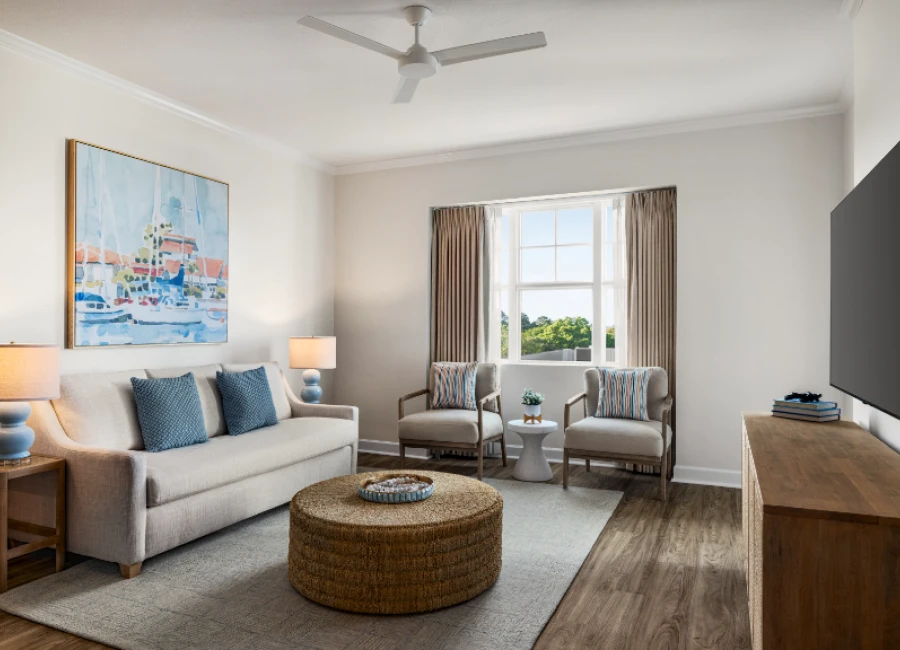 A cozy living room with a beige sofa, blue pillows, two matching armchairs, a woven ottoman, side tables with lamps, a large painting, a window with curtains, and a mounted TV across from a wooden console.