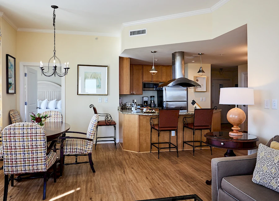 A bright, open living-dining area with a kitchen bar, cozy sofa, patterned chairs, a fireplace chandelier, and warm wood flooring.