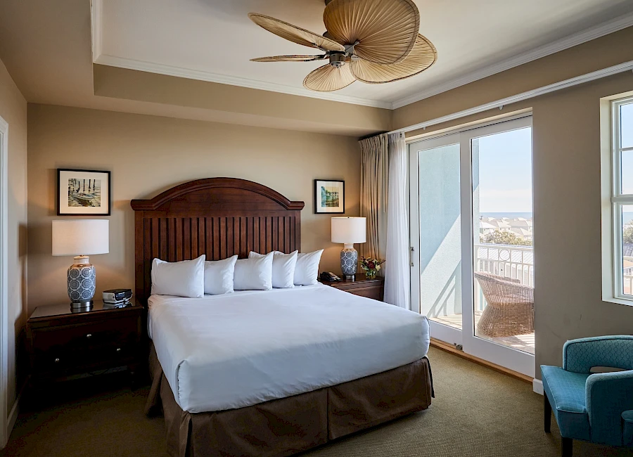 A tidy hotel bedroom with a large bed, two nightstands, lamps, and a seating area by large windows offering a balcony view.