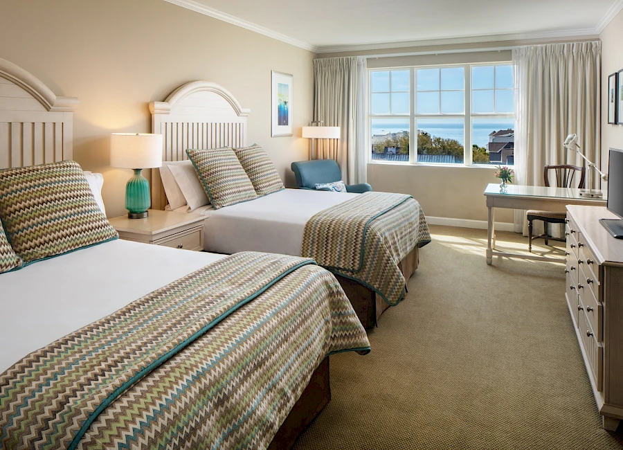 A bright hotel room with two queen beds, neutral decor, nightstands, lamps, a desk by the window, and a view of the ocean through the bay window.
