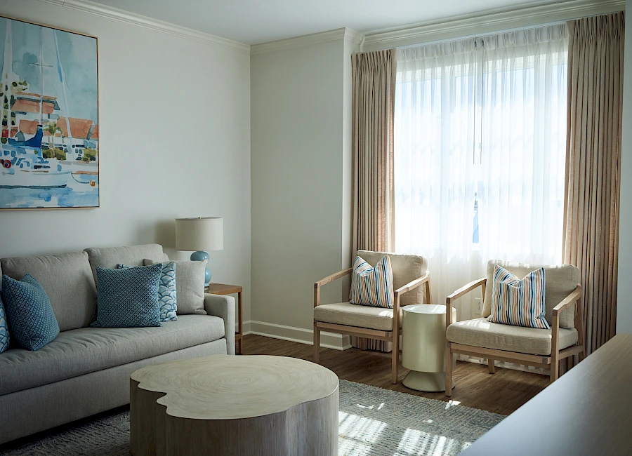 A bright living room with a gray sofa, blue throw pillows, two wooden armchairs by a window, a round coffee table, and soft natural light.