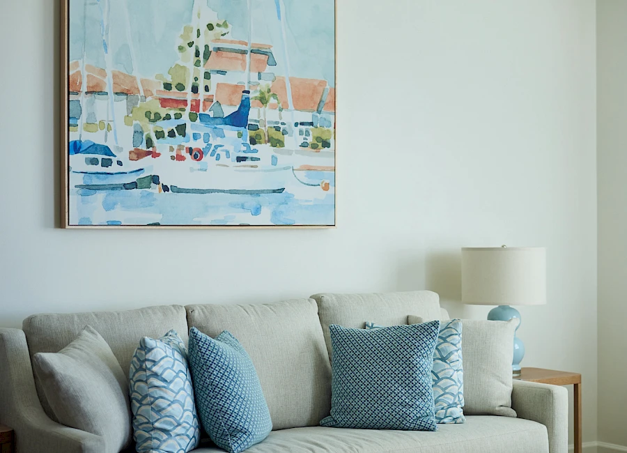 A cozy living room with a light grey sofa, blue patterned throw pillows, a round wooden coffee table, a harbor painting, and a white lamp.