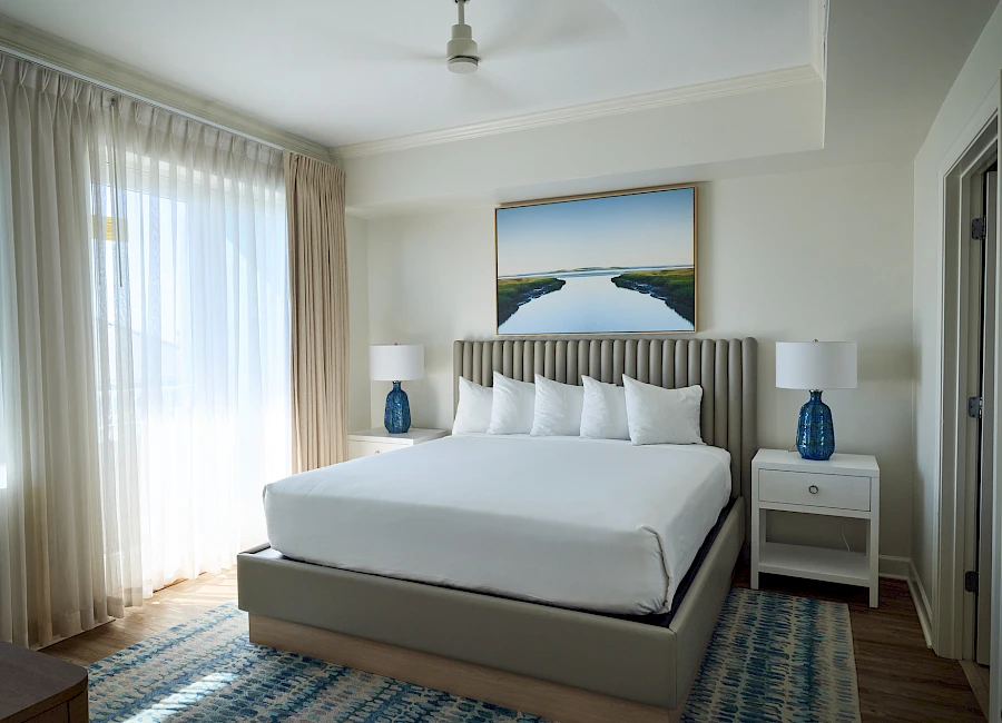 A bright hotel-style bedroom with a large bed, white bedding, two matching nightstands with blue lamps, sheer curtains, and a coastal painting above the headboard. The room has a light, airy feel with a patterned blue rug.
