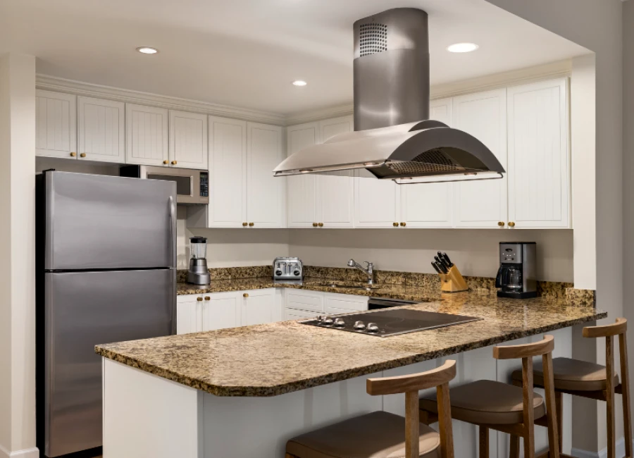 A modern kitchen with white cabinets, granite countertops, stainless steel appliances, and a central island with three stools under a stainless range hood.