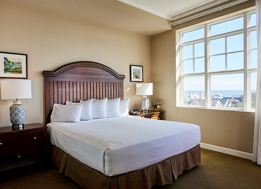 A neatly made hotel room with a large wooden headboard, two nightstands, lamps, white bedding, and a big window offering a city view.