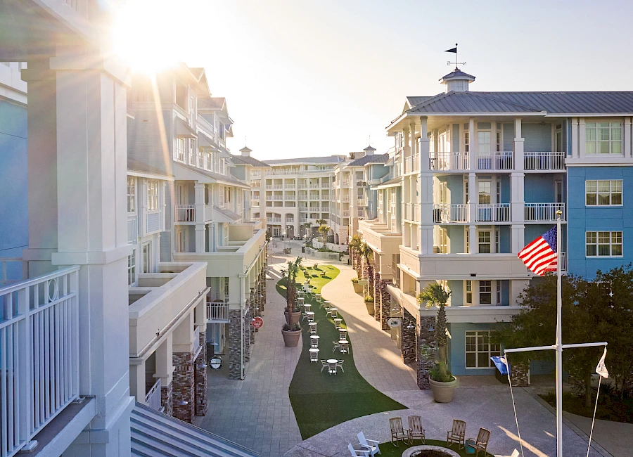 A sunny coastal resort with pastel condo buildings lining a central courtyard path, balconies, palm trees, and flags waving by the dunes.