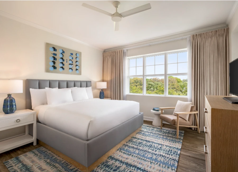 A bright hotel room with a plush gray bed, white linens, two nightstands, blue vases, a ceiling fan, large window with beige curtains, and a cozy seating area by the view, all ending with a period.