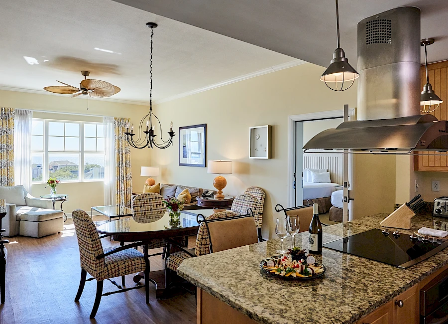 A bright, open kitchen-dining area with a granite island, hanging lights, a round table, and a view of a cozy living space.