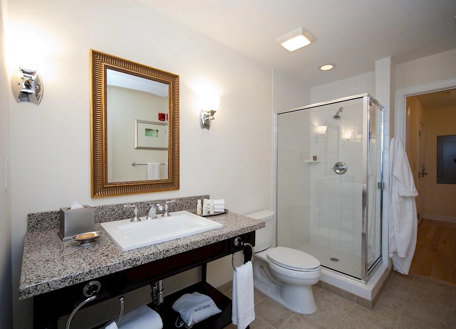 A modern hotel bathroom with a granite counter, sink, framed mirror, wall sconces, a glass-enclosed shower, toilet, and towels under the counter, all bright and clean.
