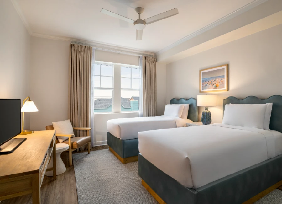 A bright hotel room with two twin beds, blue upholstered headboards, a desk with a lamp, a chair, a window with beige curtains, and soft decor.