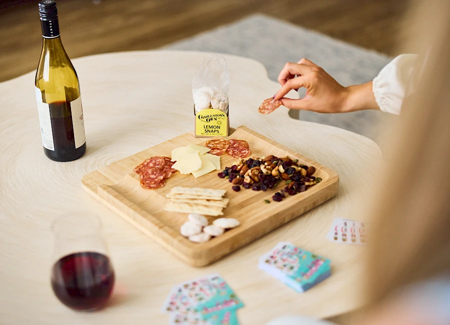 A woman prepares a cheese and charcuterie board with crackers, nuts, and grapes, while a bottle of wine, a glass, and playing cards sit nearby.