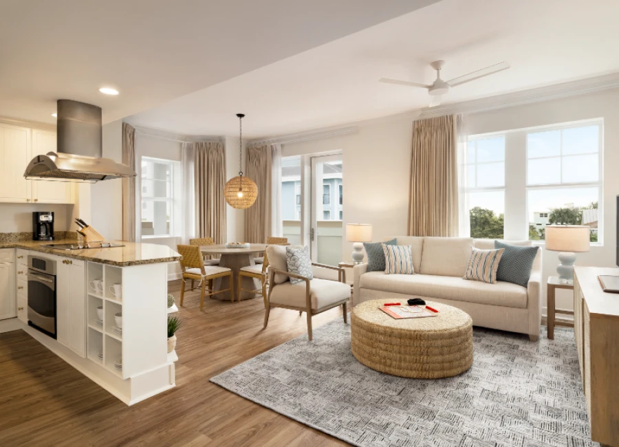 A bright, open-concept living space with a cozy gray rug, beige sofa, round woven coffee table, dining area, and a modern kitchen nearby with white cabinets and stainless appliances.